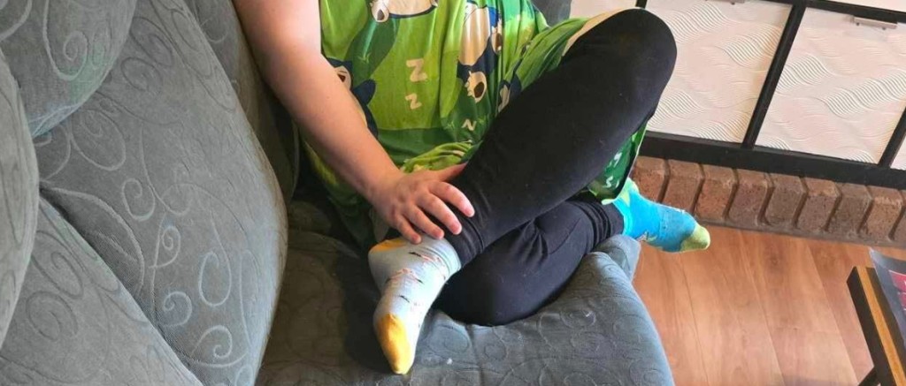 Femme assise sur canapé en tenant ses jambes relevées dans une position atypique.